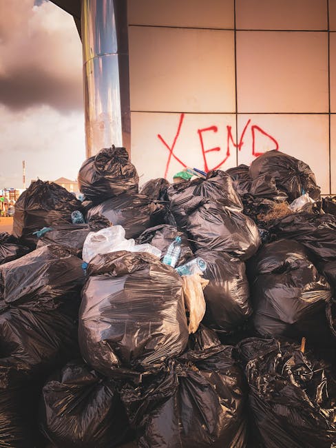 A large pile of black garbage bags, some torn open, and empty plastic bottles scattered among them, situated against a modern building with a tiled white facade and red graffiti reading 'XEND' on the wall behind. The bags are stacked on a paved surface, likely a street or alleyway, with a cloudy sky visible in the background. The scene appears to be an outdoor area where refuse has been collected or left for disposal. The overall impression highlights the volume of waste associated with private rubbish collection and the importance of professional rubbish removal services like Rubbish Removal Earls Court to manage such waste efficiently and responsibly.