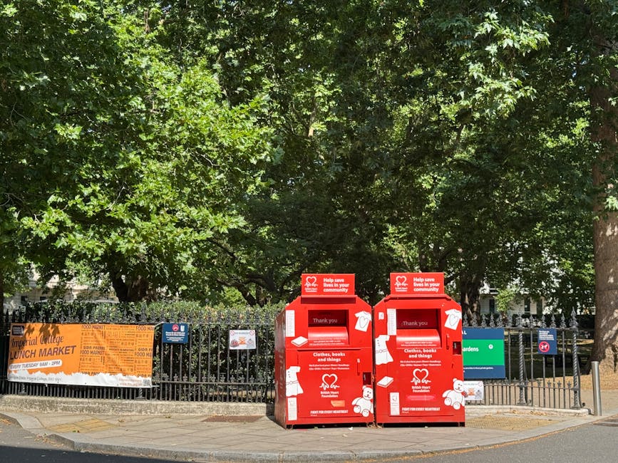 Two bright red charity collection bins with white and red signage are positioned on a paved sidewalk near a black metal fence. The bins are labeled for collecting clothes, books, and miscellaneous items, with printed icons of clothing and books. The bins feature a slot at the top for inserting donations and a small window on the side. Behind the fence, a green space with dense foliage of trees and bushes is visible, creating a lush, leafy background. A large orange banner advertising a lunch market is attached to the fence on the left side, with partial text visible. The scene is illuminated by natural daylight, casting soft shadows and highlighting the vivid colors of the bins and the greenery. The setting appears to be in an urban area with a mix of natural elements and community infrastructure, aligning with sustainable waste handling or alternative disposal options, as seen with private charity collection points, which complements the context of rubbish removal services handled by Rubbish Removal Earls Court.