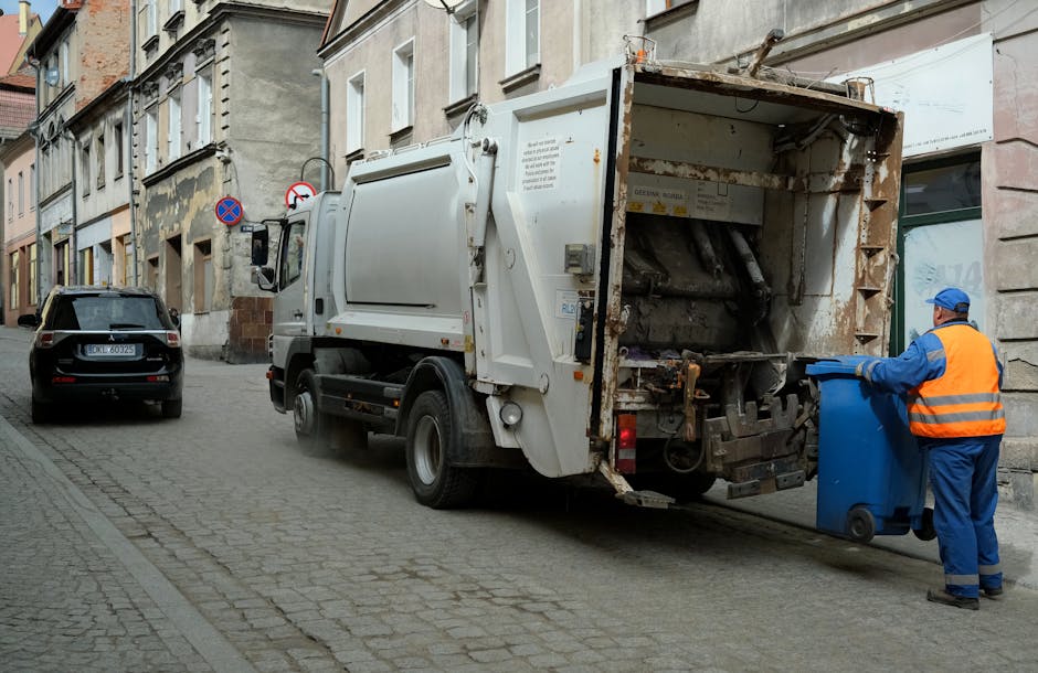A street scene in an urban residential area showing a large white refuse collection vehicle parked on a cobblestone road with its rear compactor open, revealing the rusted, dark grey metal interior used for compacting waste. To the right, a worker dressed in a blue uniform and orange high-visibility vest is disposing of rubbish into a blue wheeled bin. The background includes multi-story buildings with a mix of beige and grey facades, some showing signs of age and weathering, with narrow windows and decorative architectural features. A black car is parked further along the street on the left side, and two traffic signs indicating parking restrictions are mounted on a pole attached to a building wall. The overall scene conveys a typical rubbish collection operation in a densely built area, aligning with private waste disposal services like those offered by Rubbish Removal Earls Court.