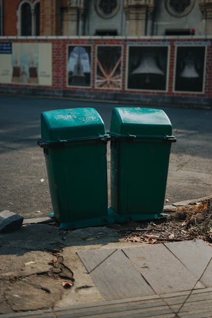 Two bright red charity collection bins with white and red signage are positioned on a paved sidewalk near a black metal fence. The bins are labeled for collecting clothes, books, and miscellaneous items, with printed icons of clothing and books. The bins feature a slot at the top for inserting donations and a small window on the side. Behind the fence, a green space with dense foliage of trees and bushes is visible, creating a lush, leafy background. A large orange banner advertising a lunch market is attached to the fence on the left side, with partial text visible. The scene is illuminated by natural daylight, casting soft shadows and highlighting the vivid colors of the bins and the greenery. The setting appears to be in an urban area with a mix of natural elements and community infrastructure, aligning with sustainable waste handling or alternative disposal options, as seen with private charity collection points, which complements the context of rubbish removal services handled by Rubbish Removal Earls Court.