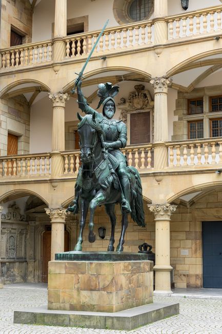 A detailed bronze equestrian statue depicting a medieval knight in armor, mounted on a sturdy stone plinth in an outdoor courtyard area. The knight is shown wearing a helmet with a crest, a chainmail or chainmail-like armor, and a tabard or surcoat draped over the armor. The knight holds a sword raised overhead in his right hand, with the blade pointing upward, while his left hand rests on the reins of the horse. The horse is depicted with a bridle and saddle, standing firmly on all four legs. The surrounding environment features an ornate building with classical architectural elements, including a curved balcony with balustrades, supporting columns with decorative capitals, and warm-toned stone walls. Behind the statue, there are windows and entrances, with the stonework exhibiting a weathered, textured appearance. The scene has soft, natural lighting, highlighting the details of the bronze sculpture and the building's facade, creating an overall impression of historical significance and craftsmanship related to the theme of cultural heritage and civic pride. Rubbish removal services at Rubbish Removal Earls Court can manage waste from such historic or civic sites, providing independent collection options for careful site maintenance and preservation.