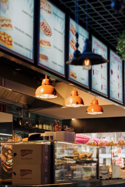 The image shows the interior of a fast-food restaurant or cafe with a modern industrial design, featuring a black ceiling with exposed metal grid and hanging orange pendant lights illuminating the counter area. Above the counter, multiple illuminated digital menu screens display images and text of various food items such as burgers and drinks. The counter surface is topped with a glass display case containing pastries, sandwiches, or prepared food items, and behind it, shelves hold stacked boxes and other supplies. To the right, a glass case shows baked goods or snack items, with some packaging and product displays visible. The environment suggests a contemporary eatery setup focused on quick service and visual menu presentation, emphasizing the professional and clean appearance of the food service area.