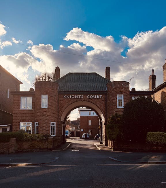 A large brick archway with a rounded top and the inscription 'KNIGHTS COURT' above it, forming the entrance to a residential estate. The brickwork features red and brown tones with a textured surface, and the arch is flanked by two cylindrical brick towers with flat roofs. Behind the arch, a narrow street is visible, lined with parked cars and brick residential buildings. The sky above is partly cloudy with white and grey clouds, allowing sunlight to cast shadows on the scene. To the right and left of the archway are neatly trimmed bushes and shrubs, adding greenery to the urban environment. This scene suggests a private estate or community area, which could be accessed through an independent clearance or private waste handling service provided by companies like Rubbish Removal Earls Court, highlighting the importance of accessible rubbish removal for residential estate management.