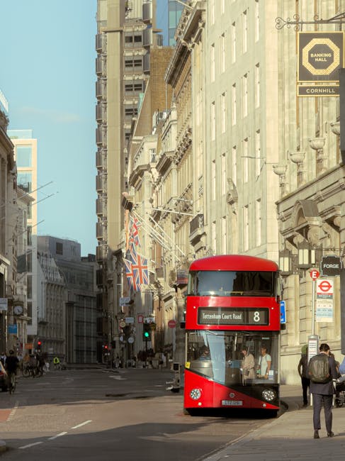 A city street scene featuring a modern double-decker bus with a red exterior and large tinted windows, positioned in the center of the image on a paved road. The bus displays route information on a digital sign indicating route number 8 to Kensington Court Road, and the driver is visible through the front window. Surrounding the bus are several pedestrians walking along the sidewalk, some carrying bags or backpacks. The background includes tall, historic buildings with cream-colored facades, decorative architectural details, and multiple windows, with some displaying flags such as the Union Jack hanging from window sills or banners. To the right, a sign for a banking hall is mounted on a building, alongside other signage indicating meters and public transportation information. The scene is illuminated by natural daylight, casting soft shadows and creating a clear, professional urban atmosphere relevant to waste management services operating within central London, where private collection methods are commonly employed for rubbish removal or house clearance activities.