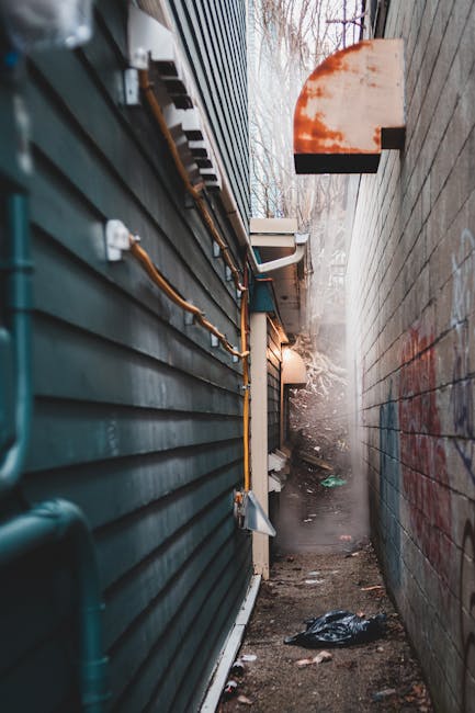 A narrow alleyway situated between two buildings, with the left side featuring dark green horizontal siding made of metal or vinyl, and the right side displaying a plain, weathered brick wall with some red graffiti marks. Along the green siding, there are visible exposed pipes and electrical wiring, with some vertical and horizontal conduits secured to the surface. The ground is composed of uneven dirt and scattered debris, including crumpled black plastic and small trash items. At the far end of the alley, a faint mist or dust is visible, partially obscuring the background which shows leafless tree branches and a cloudy sky, suggesting an outdoor environment in colder weather. An external wall-mounted light fixture with an orange-tinted semi-circular cover protrudes from the brick wall, casting a subtle glow. The scene appears to be part of a service or waste management context, possibly indicating an area used for rubbish removal or private waste collection, with the narrow space often associated with difficult access for conventional waste disposal methods. Rubbish Removal Earls Court may use such spaces for alternative waste handling solutions, given the limited access and constrained environment.