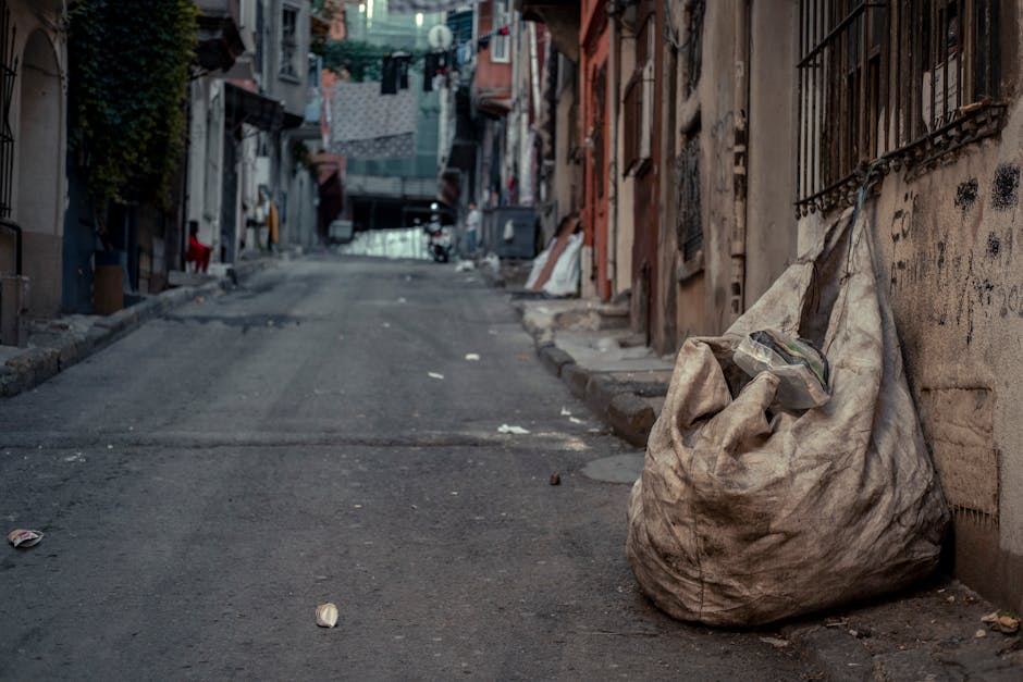 A large, tightly tied white rubbish sack rests against a weathered brick wall on the right side of a narrow, uneven urban street. The sack appears worn and stained, and some waste materials are visible protruding from the top. The street surface is rough, with small scattered pieces of litter and debris, leading into a slight incline beneath a canopy of aged buildings with metal and wooden window frames, some of which have iron bars. In the background, laundry is hanging on clotheslines strung between buildings, and the scene is illuminated by natural light, creating a muted, everyday atmosphere. This setting suggests an informal or independent rubbish collection context, potentially supported by services like Rubbish Removal Earls Court, prioritizing alternative waste handling in a densely built residential area.